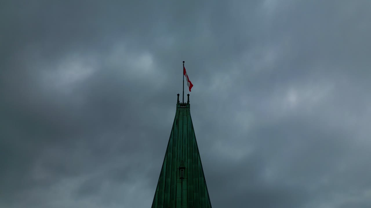 Zbliżenie z drona wieży zegarowej Ottawa Hill, przedstawiające Kanadyjską Flagę na tle zachmurzonego letniego nieba i schodzące do zegara, oddające atmosferę ponurości i politycznego niepokoju.