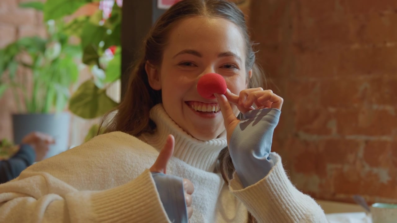 Close up of young woman holding and adjusting her red clown nose in indoor setting. Woman wears white turtleneck sweater with soft lighting highlighting texture of fabric against warm brick