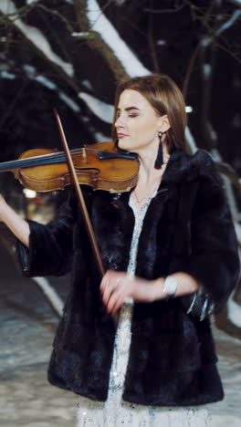 a beautiful girl with make-up and in a white dress is playing a violin composition in a winter park on the background of snow. camera motion to left vertical video