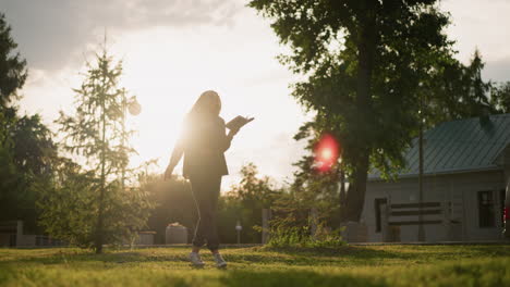 dama de ropa gris bailando alegremente al aire libre en un campo cubierto de hierba con un libro en la mano, mientras la luz del sol crea una atmósfera radiante a su alrededor, con edificios y vegetación en el fondo