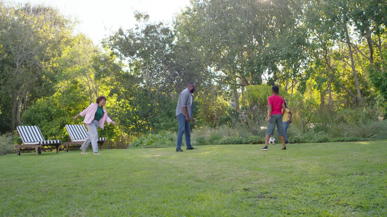 African American family playing soccer on lawn, soccer ball rolling, youth in yellow retrieving