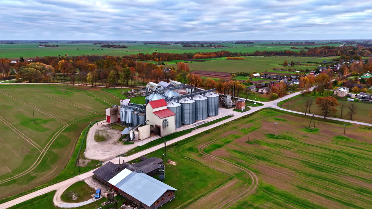 Ripresa aerea con effetto parallasse di silos per lo stoccaggio del grano e impianto di sollevamento circondati da campi verdi e alberi autunnali nella campagna rurale.