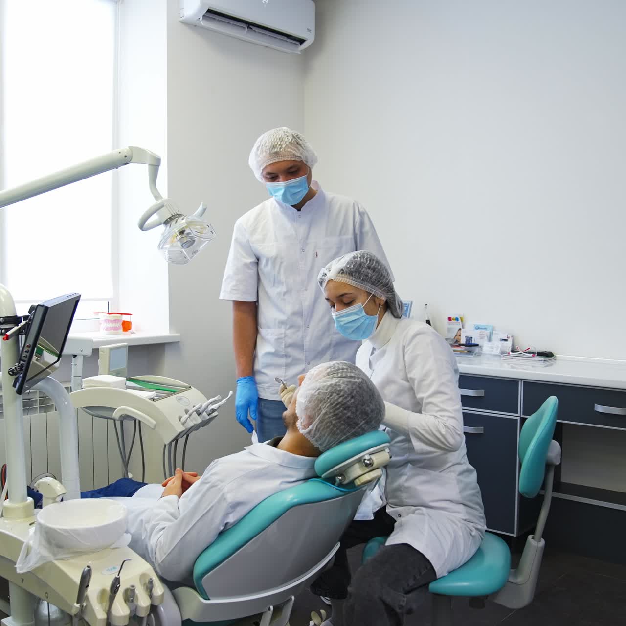 Practical lessons at dentistry department of university. Female student takes metal tool from a tray for examination of a patient. Male students stands behind and assists