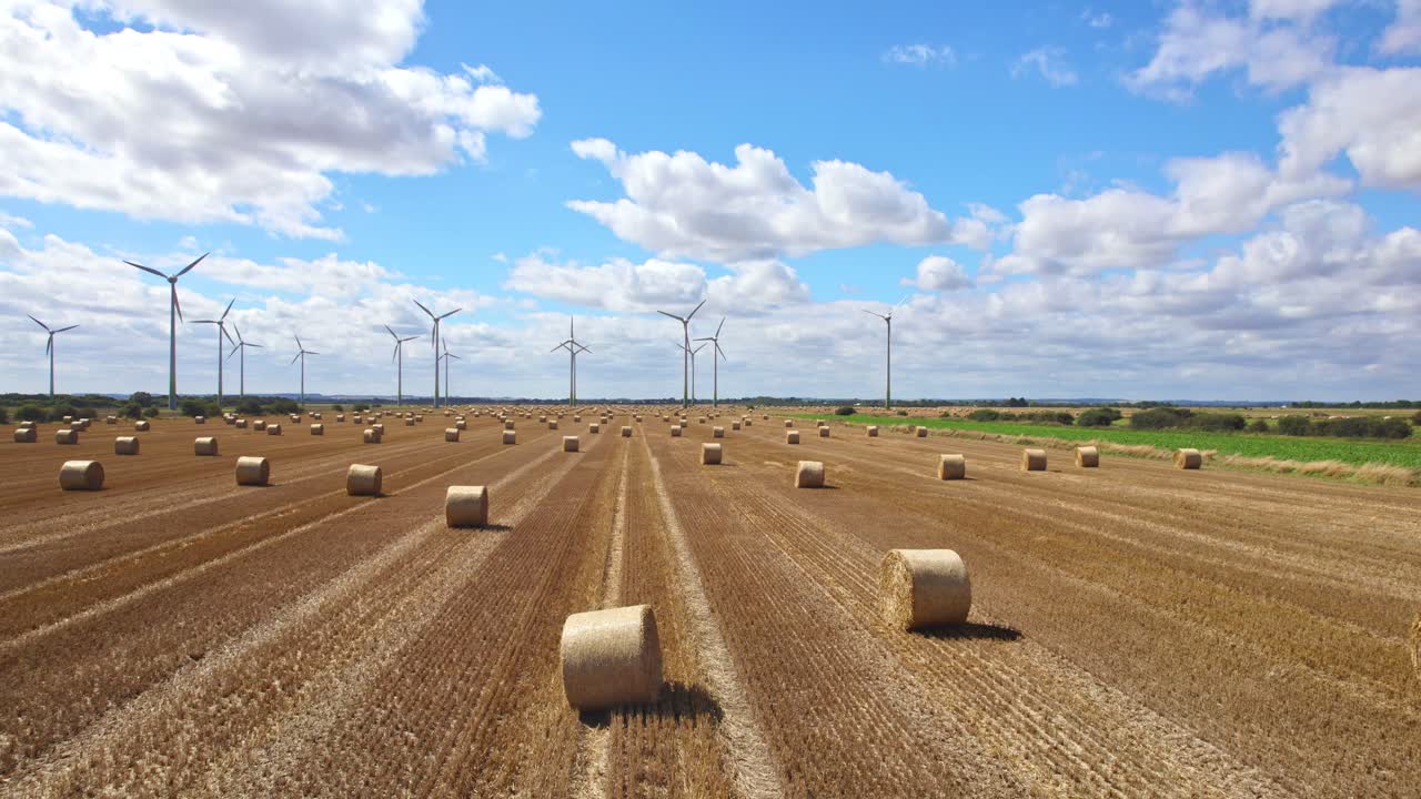 An aerial video showcases the beauty of wind turbines in a row, spinning elegantly within a Lincolnshire farmer's just-harvested field, accented by golden hay bales