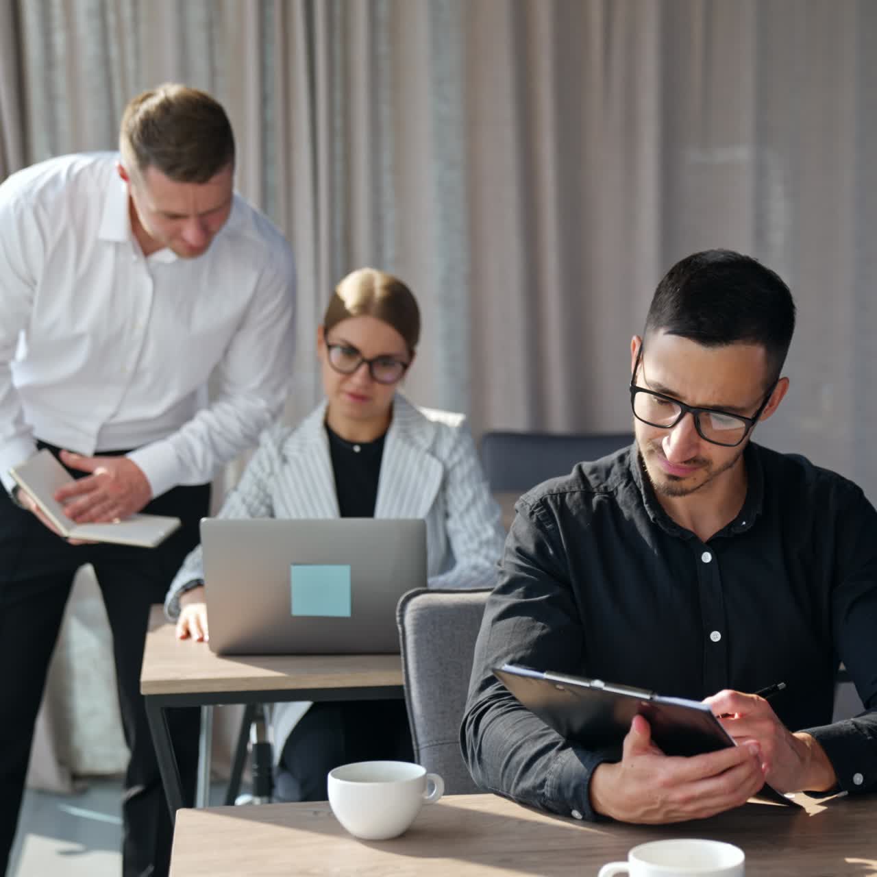 Man in black shirt and glasses checking information in papers and on computer screen. Male and female colleagues at desk behind consulting looking at laptop