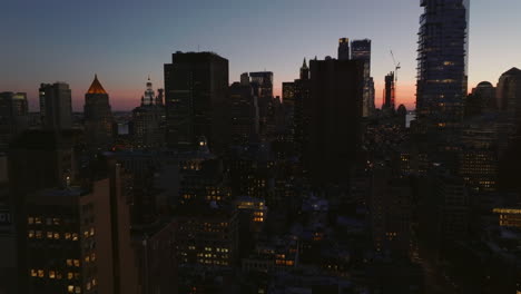 Fly-above-city-at-dusk.-High-rise-buildings-with-lighted-windows.-Silhouettes-against-colourful-sky.-Manhattan,-New-York-City,-USA