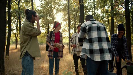 A-happy-group-of-people-on-a-hike-stop-at-a-halt-and-take-off-their-shoulders-their-backpacks-in-the-summer-forest.-A-brunette-man-in-a-green-jacket-communicates-with-his-friends-on-a-hike-and-tells-what-everyone-needs-to-do.-Distribution-of-responsibilities-during-the-trip