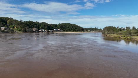 a steady camera captures a broad river under blue skies, with gentle water movement, lush green banks, and distant houses in natural daylight