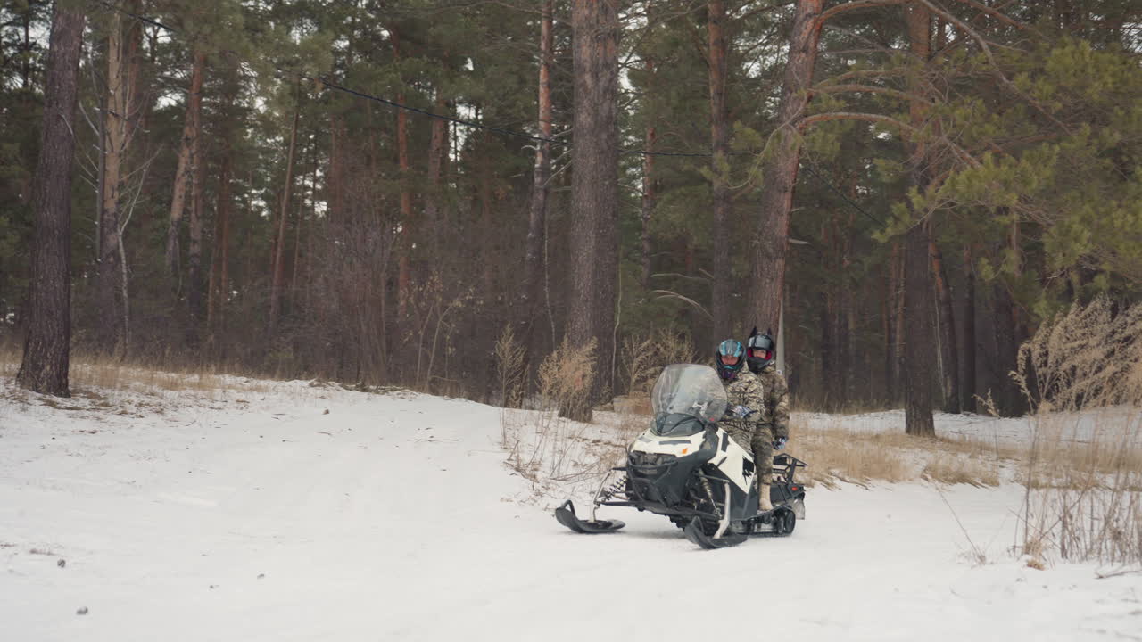 Due piloti su motoslitta in una foresta, con attrezzatura mimetica e caschi, si avvicinano a una radura sotto la neve che cade. Si sentono il rombo del motore e le tracce lasciate. Una stretta collaborazione e un controllo condiviso, lavoro di squadra su un sentiero ghiacciato, in un inverno rigido.