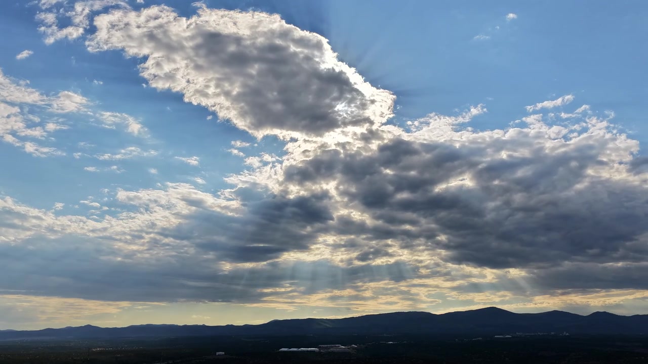 A 4k drone timelapse of hazy sunset rays shining through clouds over Los Alamos New Mexico, casting bright sunbeams down onto the dark silhouetted mountain range below.