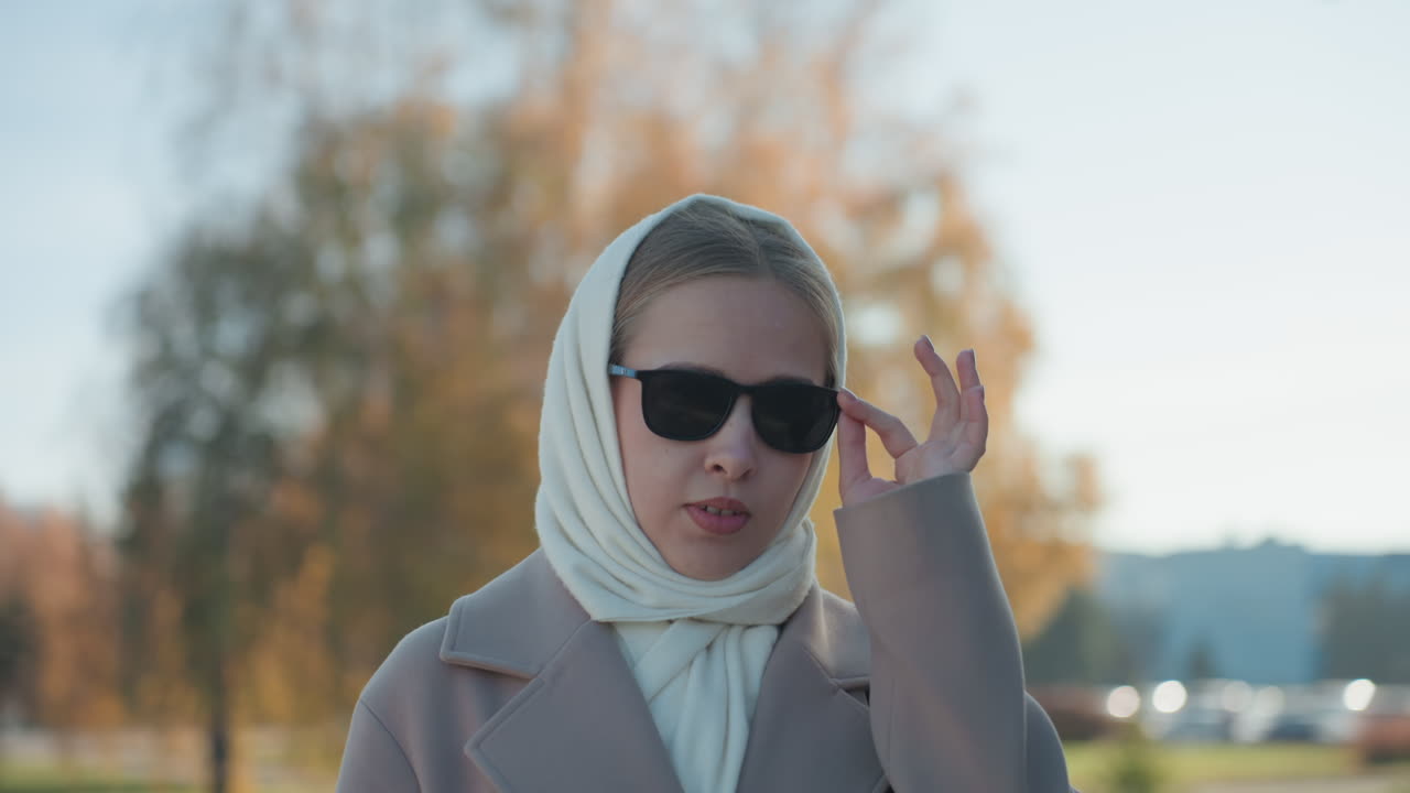 Donna in piedi tra gli alberi di un parco autunnale, ritratto cinematografico malinconico di una viaggiatrice, reporter, narratrice, con sciarpa di maglia, cappotto oversize, occhiali da sole, sguardo riflessivo, foglie cadute e luce solare soffusa.