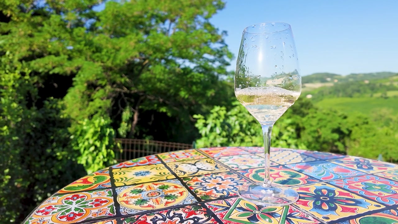 Un bicchiere di vino bianco è appoggiato su un tavolo a mosaico colorato con dolci colline verdi