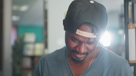Close up of man wearing backward cap, head bowed with warm smile, focusing intently on something, as bright light softly illuminates background with blurred bookshelves