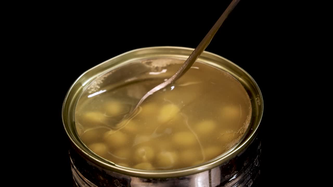A metal spoon stirs and lifts green peas from an open can filled with liquid, under bright studio lighting with a black background