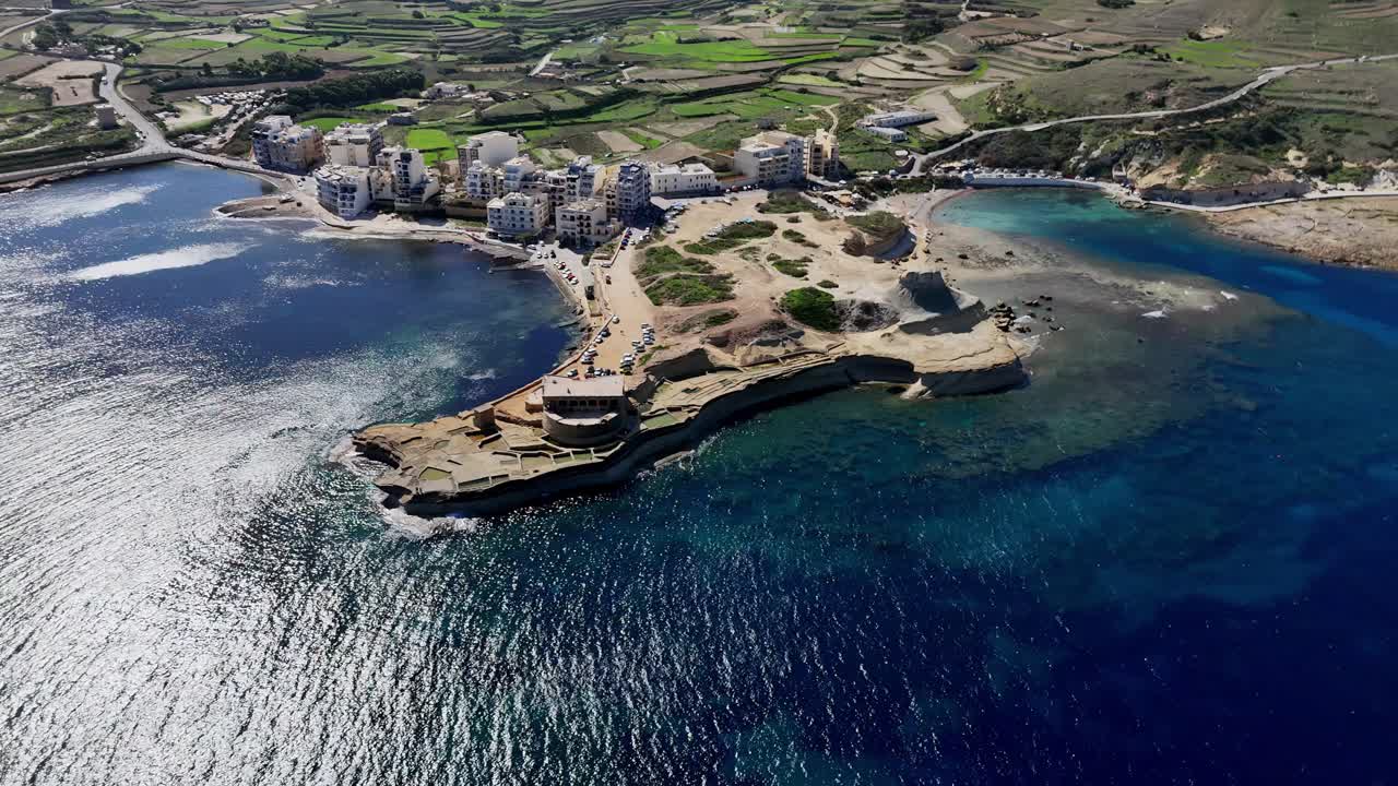 Panoraamakuva ilmasta Qbajjarin lahdesta Marsalfornissa, Gozolla, kirkkaana aurinkoisena kesäpäivänä. Näkymässä kalkkikivikallioita ja turkoosia Välimeren vettä kirkkaan sinisen taivaan alla, kuvattuna korkealta dronella.