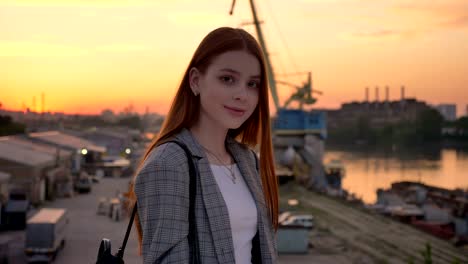 young ginger woman in jacket standing on bridge and looking in camera, industrial factory background, sunset