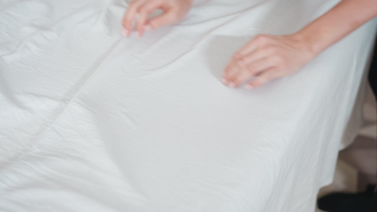 Closeup of light skin woman wearing black trousers arranging white sheet on massage bed, gently smoothing fabric with hands in calm setting, focusing on neatness and clean presentation of surface