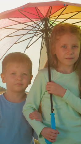 siblings huddle beneath colorful umbrella. charming brother and sister find solace under vibrant multicolored parasol enjoying cozy shelter by riverside