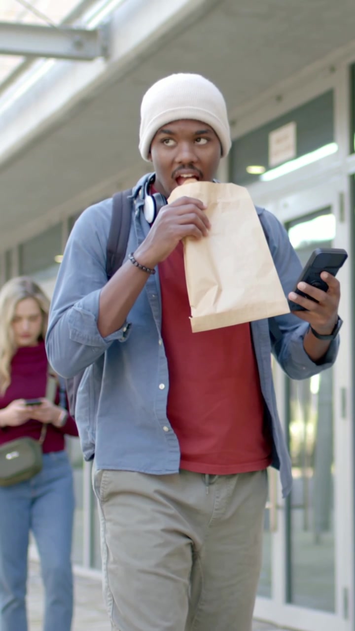 Vertical video: Walking man in beanie grabbing bag, eating pastry, checking phone on sidewalk