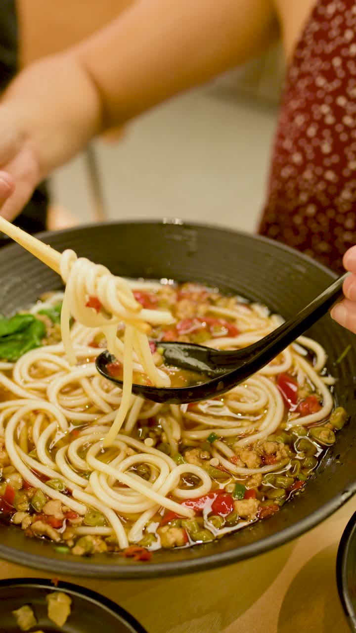 Una mano solleva noodles spessi da un brodo piccante con le bacchette, illuminazione interna calda, primo piano.