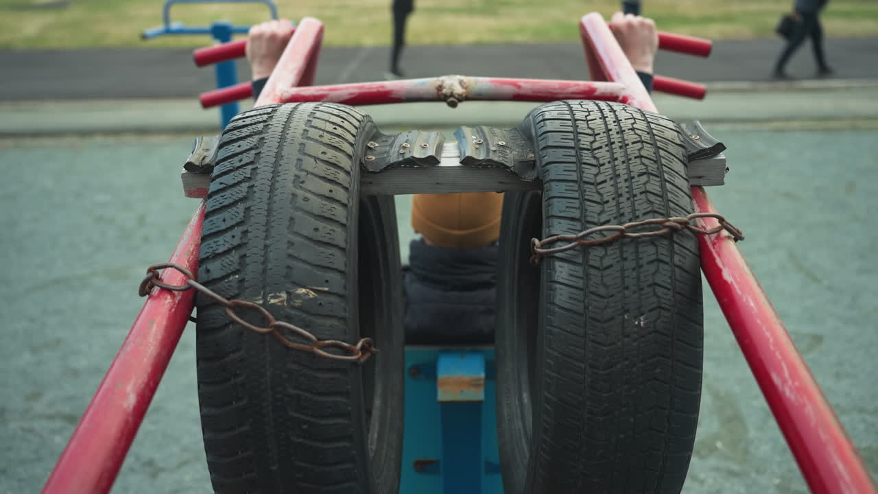 Back view of a coach doing pull-ups on workout equipment, with tires attached to bars. He pauses to move his neck side to side, with four people in the background, including two walking