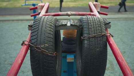 back view of a coach doing pull-ups on workout equipment, with tires attached to bars. he pauses to move his neck side to side, with four people in the background, including two walking
