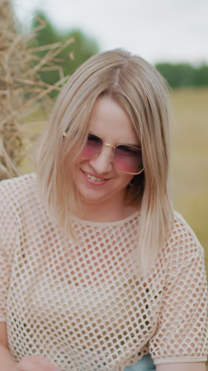 mujer con tela de red y gafas de sol sonriendo a alguien que sostiene un libro de portada marrón, con una bala de heno en el fondo y un campo abierto que se extiende en la distancia