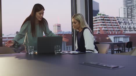 video of two diverse female colleagues discussing and using laptop in office