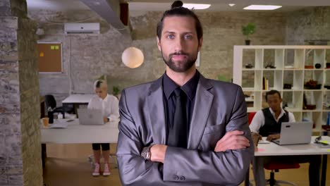 businessman with beard is standing with arms crossed and watching at camera in office, colleagues are working with laptops, work concept