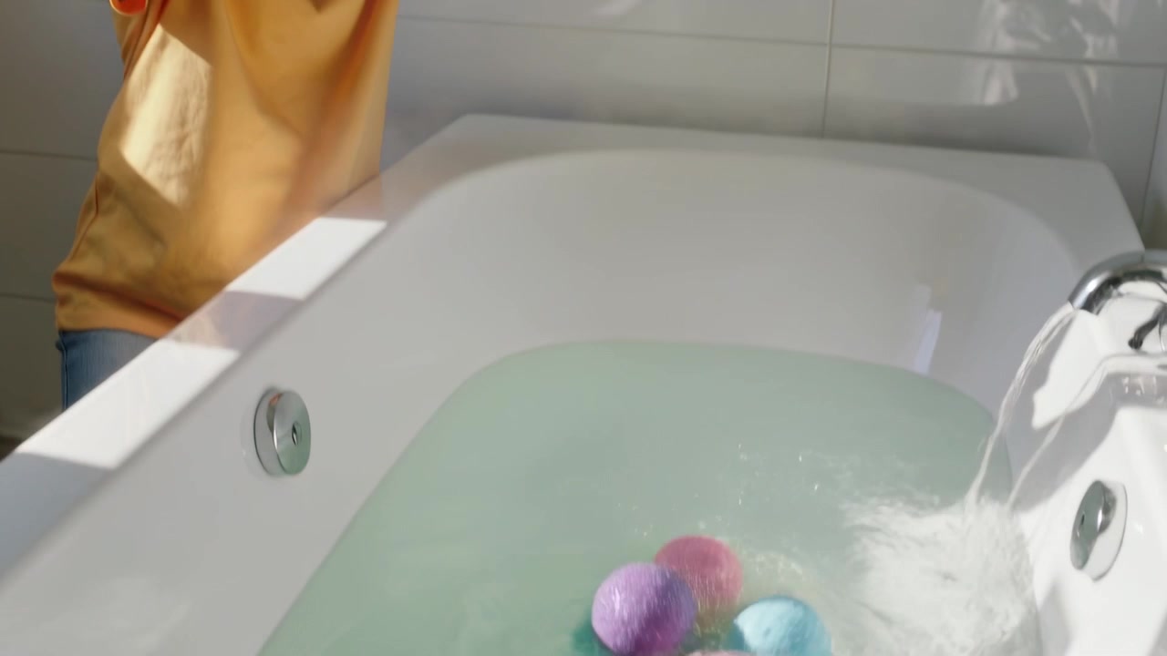 Adolescente in maglietta gialla che lascia cadere bombe da bagno pastello che colorano la vasca, rubinetto aperto, spazio per il testo.