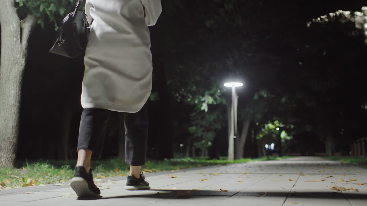Streetlight casts glow on woman in white coat walking alone along empty park path at night, surrounded by trees and shadows, her silhouette softly illuminated in peaceful solitude