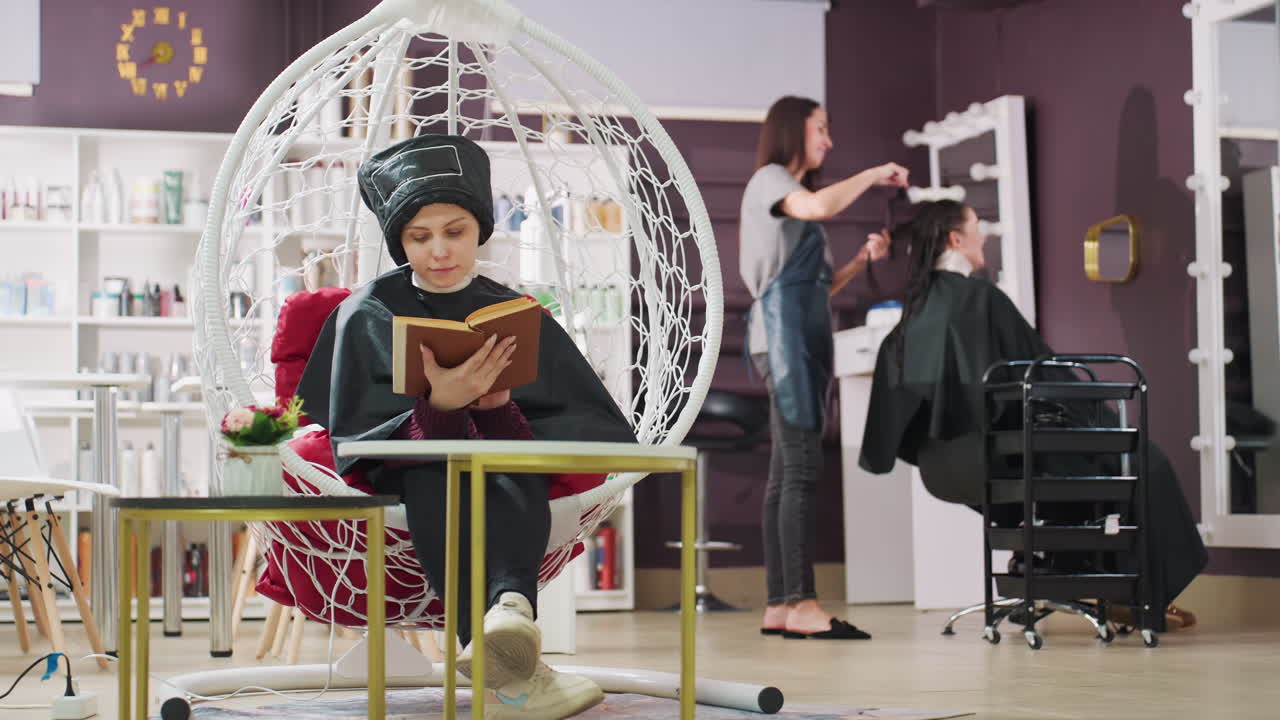 후드 드라이어 아래 살롱 의자에 앉아 책을 읽고 있는 백인 여성, 배경에는 다른 손님을 시술하는 헤어 스타일리스트, 따뜻한 조명, 플로럴 테이블, 모던한 부티크 장식, 평온한 휴식과 슬로우 라이프 분위기