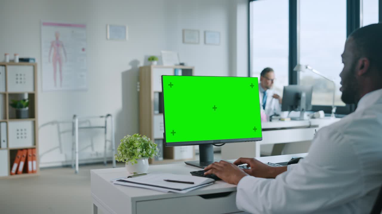 African American Medical Doctor is Working on a Computer with Green Screen Mock Up Display in a Health Clinic. Assistant in White Lab Coat is Reading Medical History Behind a Desk in Hospital Office.