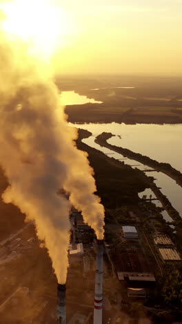 industrial factory near the river. top view of thick smoke coming from pipes into the air at sunset. manufacturing among nature. aerial view. vertical video