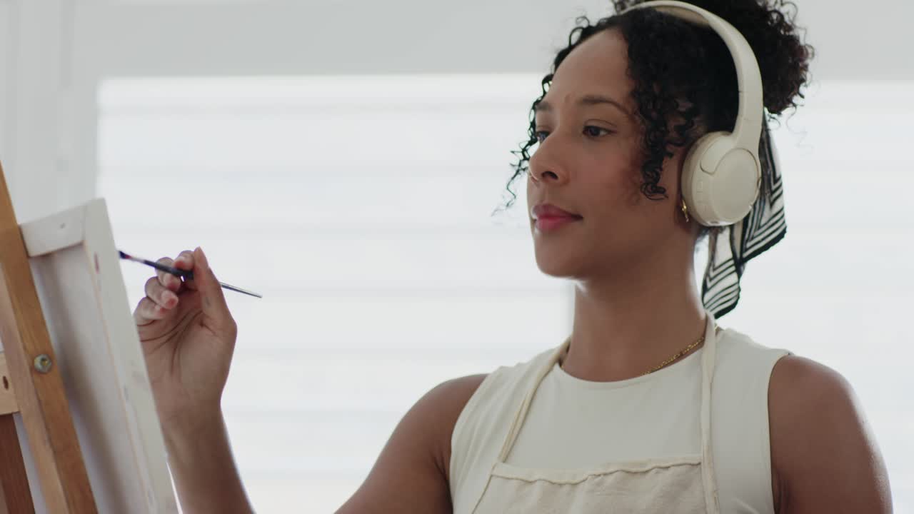 African American woman mixing paint, finishing canvas with thin brush at studio wearing headphones