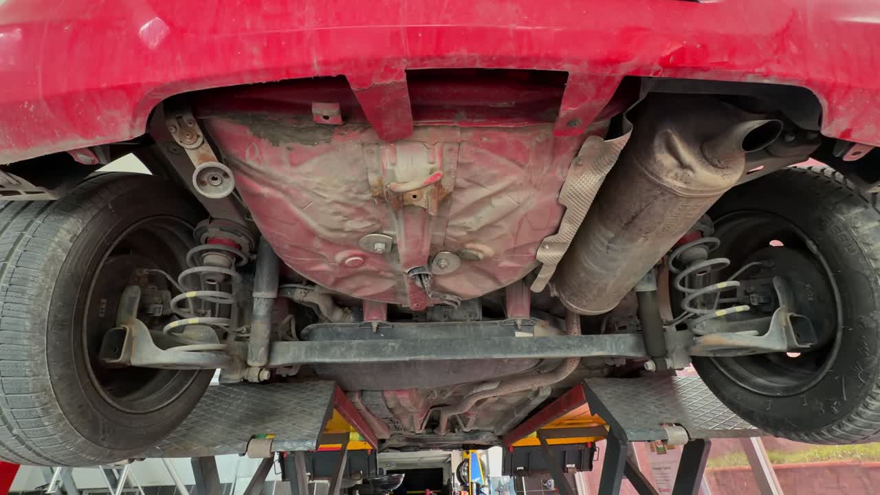 Vue du dessous d'une voiture rouge sur un pont élévateur à ciseaux hydraulique dans un atelier de réparation automobile professionnel. Montre la suspension, l'échappement et le châssis pendant l'entretien et l'inspection mécanique du véhicule.