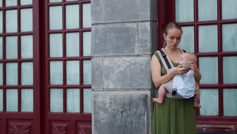 Carrying-her-infant-in-a-kangaroo-backpack,-a-young-woman-takes-photos-on-a-mobile-phone-during-her-travels.-She-walks-and-occasionally-looks-at-the-phone-screen