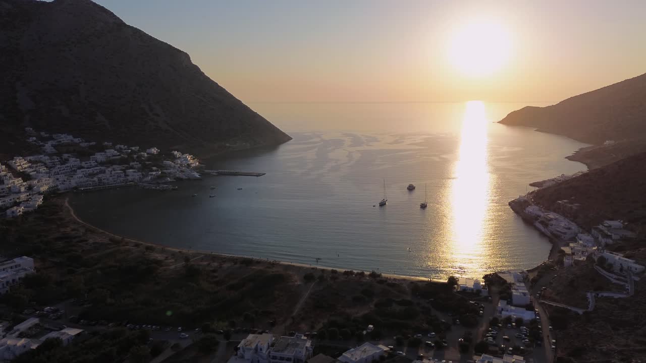 Riprese cinematografiche con drone in volo sul Mar Egeo durante il tramonto nell'ora d'oro a Kamares Sifnos, che mostrano la spiaggia sabbiosa, le case cicladiche bianche e le barche a vela ancorate nella baia.