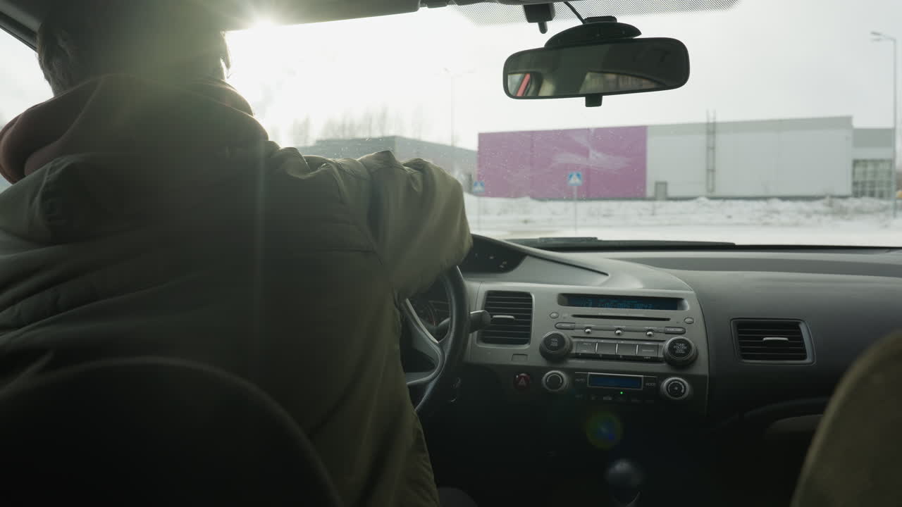 back view of man cleaning steering wheel with blue cloth inside parked car while truck and small car drive past on snowy road beyond window under soft winter light early morning calm