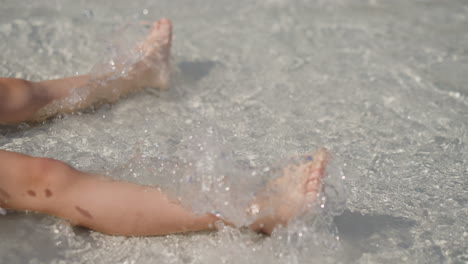 little child swings legs and splashes clear water on beach