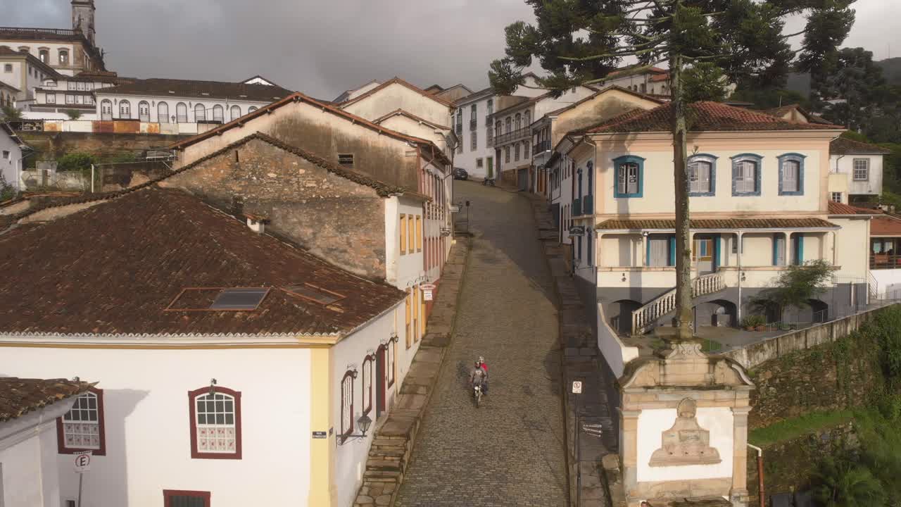 High vantage point steady view of steep cobble-street in Ouro Preto, Minas Gerais, Brazil, with a motorcycle coming down a street with typical colonial architecture