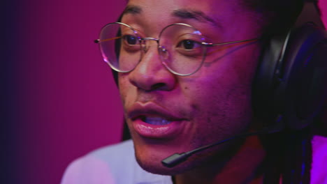 a young man wearing a headset and glasses is playing a video game. he is focused and looks excited. the background is lit with pink and blue light.