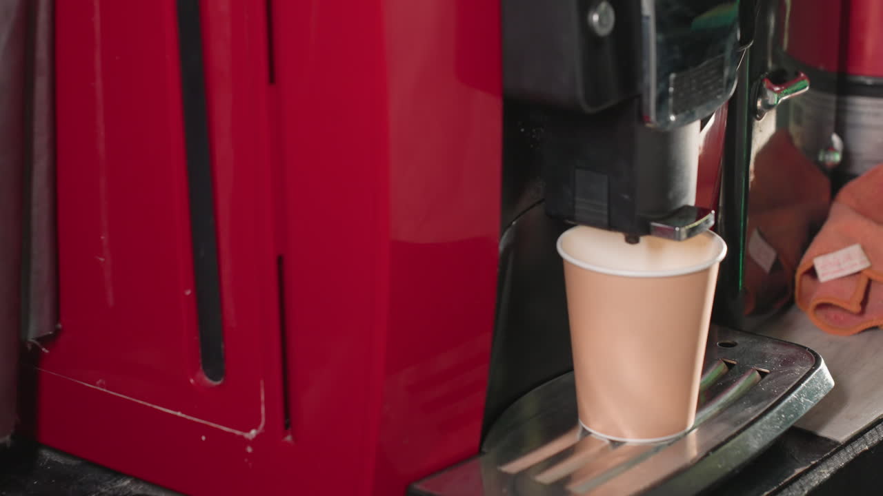Close-up shot of a coffee machine dispensing espresso into a paper cup. The machine's nozzle is visible as it releases foam or milk, emphasizing the beverage preparation process in a caf setting