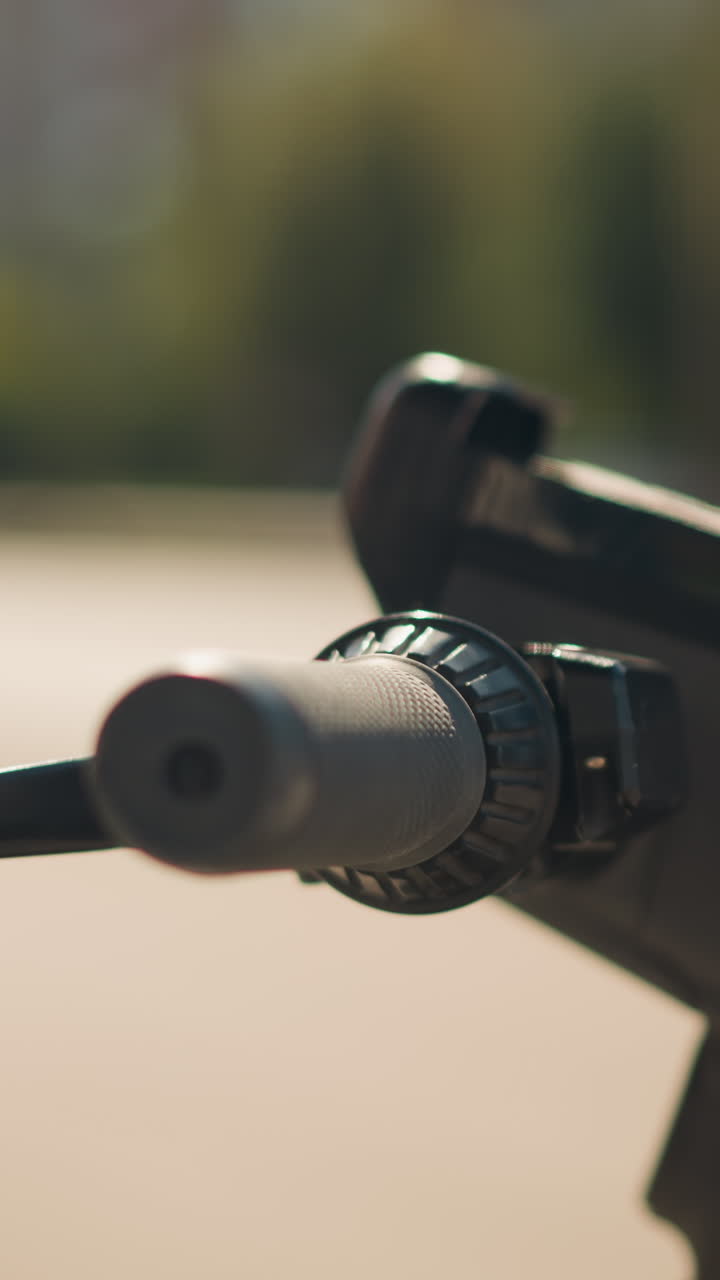 Primer plano del puño del acelerador de un patinete a la luz del sol, poca profundidad de campo que muestra la textura de goma en el manillar, el acelerador giratorio y la abrazadera de metal, fondo borroso de una plaza de la ciudad, tonos de la hora dorada.