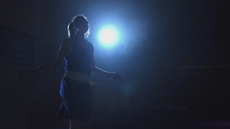 a young beautiful woman professional boxer in training in the hall jumps on a rope in the contra light in a dark room. cold light