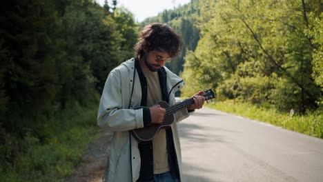 a brunette guy in a white jacket near the road plays a musical stringed instrument on a hike in the mountains