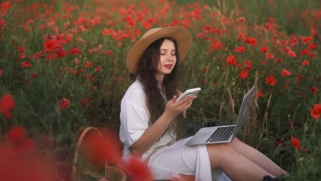 A-beautiful,-cheerful-dark-haired-girl-in-a-straw-hat,-sitting-in-a-field-of-wildflowers-and-red-poppies,-wearing-a-dress,-smiling-and-talking-on-the-phone-with-a-laptop