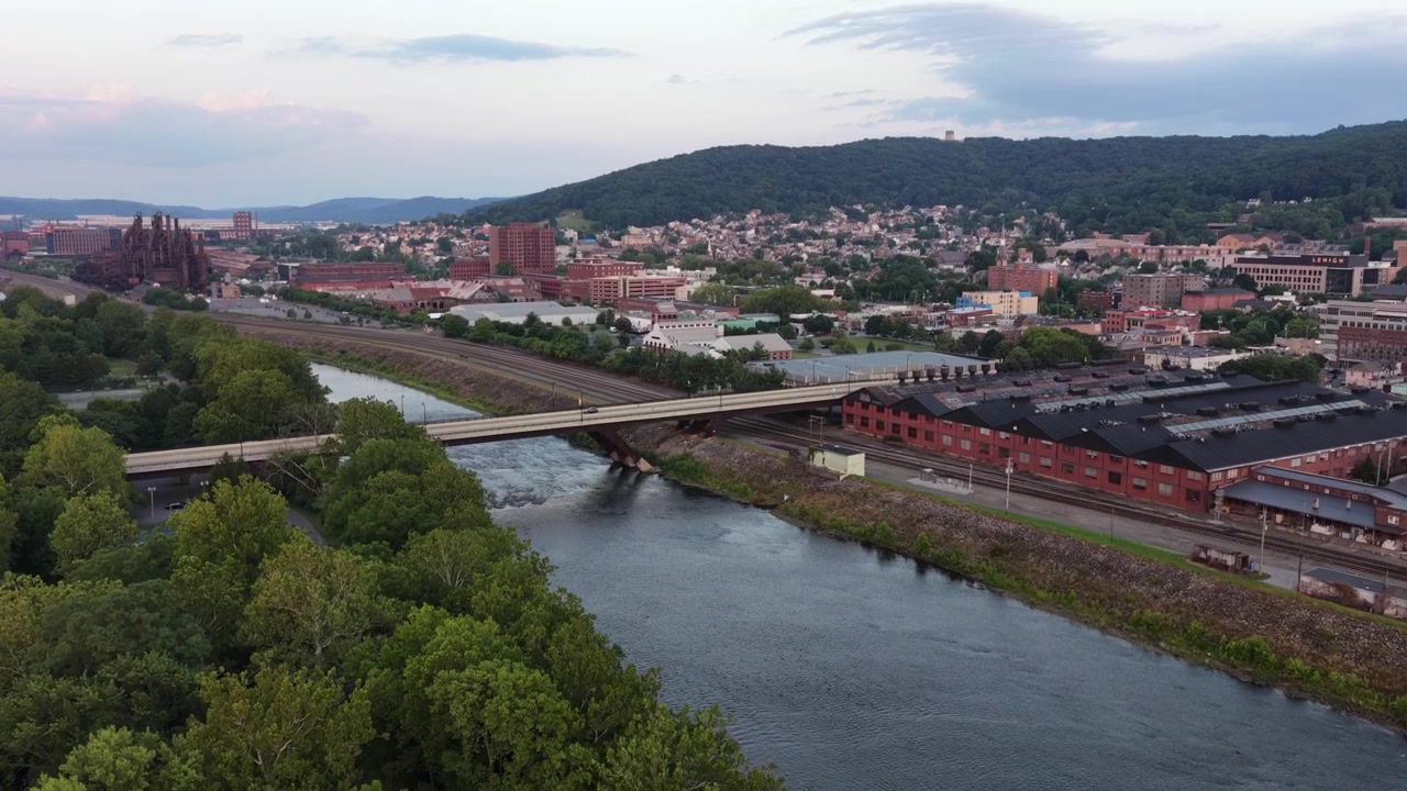 Un ponte autostradale attraversa l'ampio fiume Lehigh accanto a magazzini in mattoni rossi e binari ferroviari, con lo skyline del centro e le distanti fornaci a colata che si estendono su Bethlehem, Pennsylvania. Discesa ampia con drone aereo.