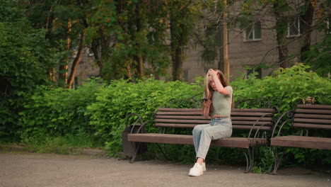 young woman sits on outdoor wooden bench, holding phone in her right hand while using her left hand to brush her hair back, surrounded by lush greenery, trees, and a building in the background