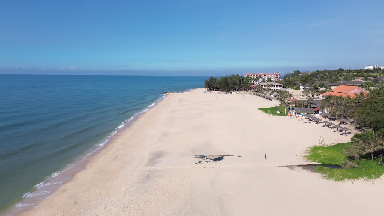 Drone flies forward over Mui Ne beach, sea left, shore centered, resorts right. Overflies someone sitting on a chair and other people walking on the white sandy shore.